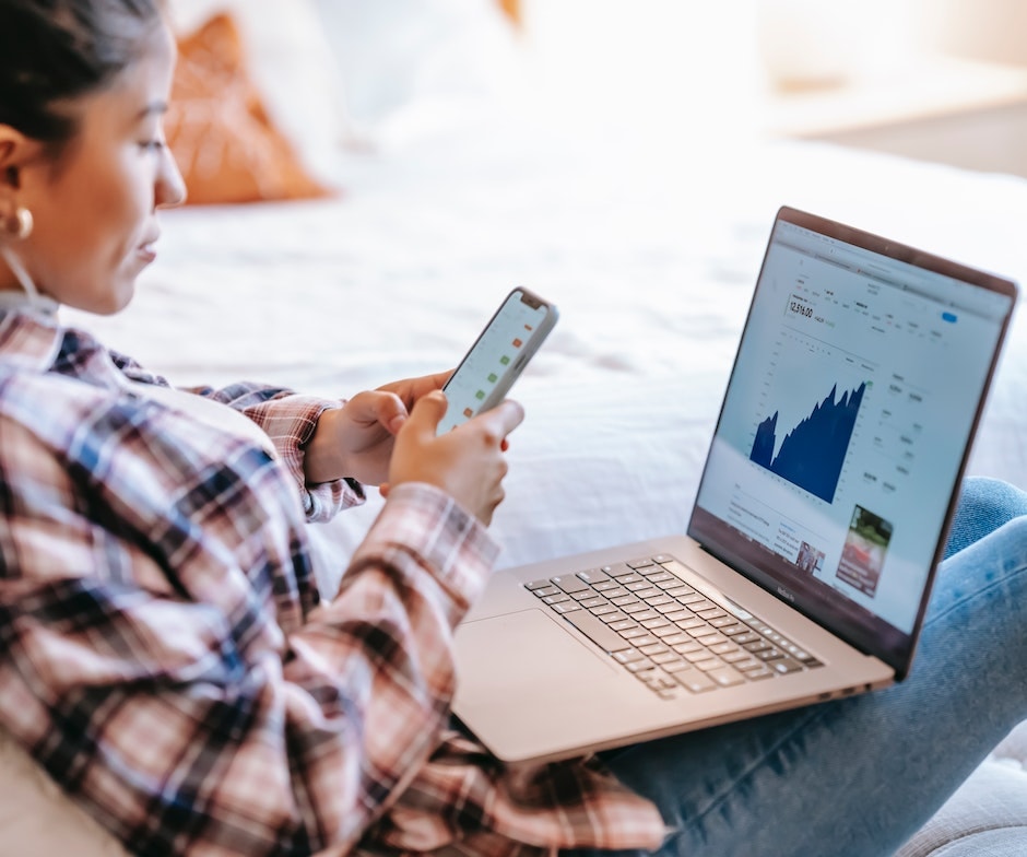 Persona sosteniendo su teléfono sentada en el sofá y un portátil con un gráfico en pantalla